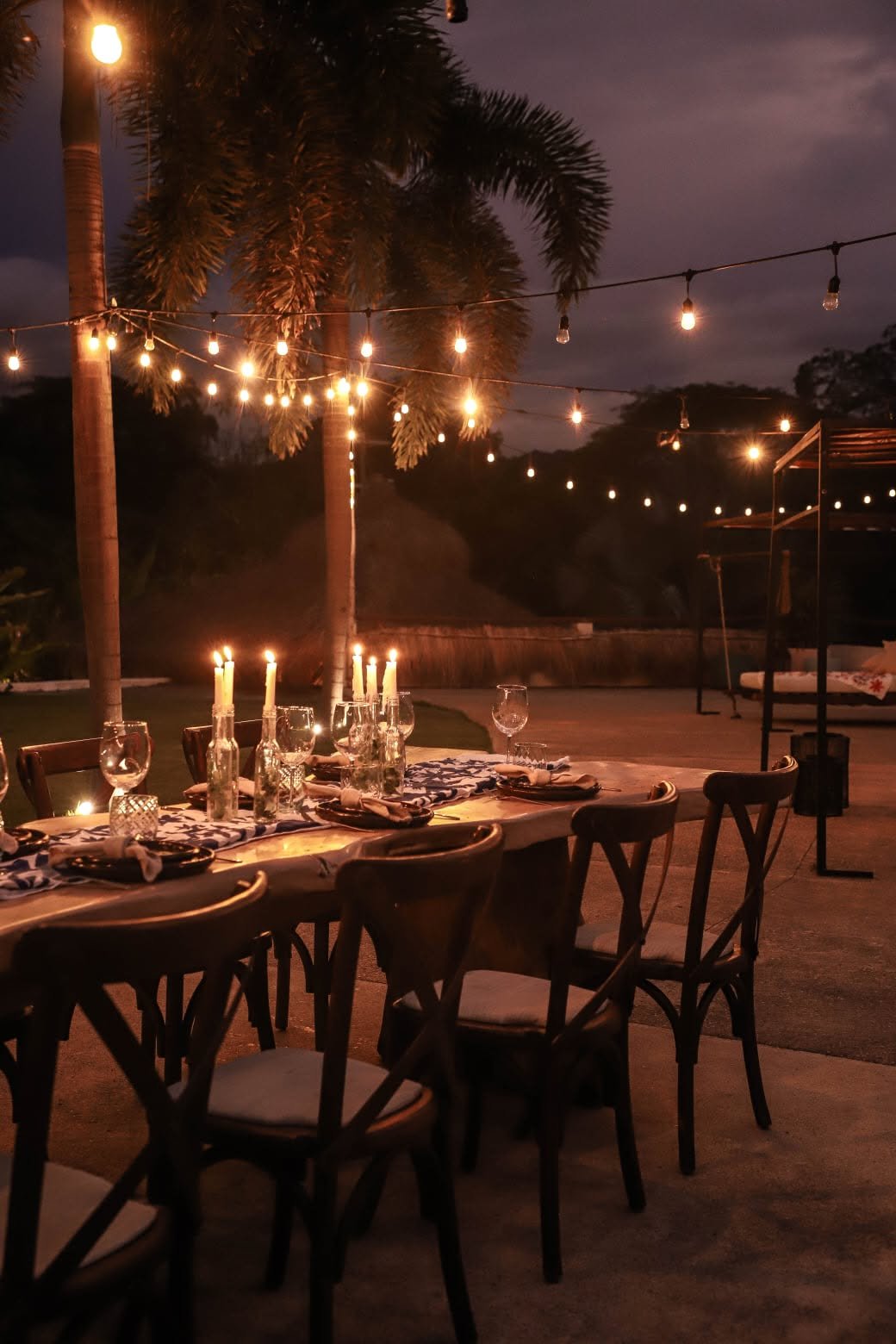 Mesa larga con velas encendidas y luces colgantes montada en el jardín de noche