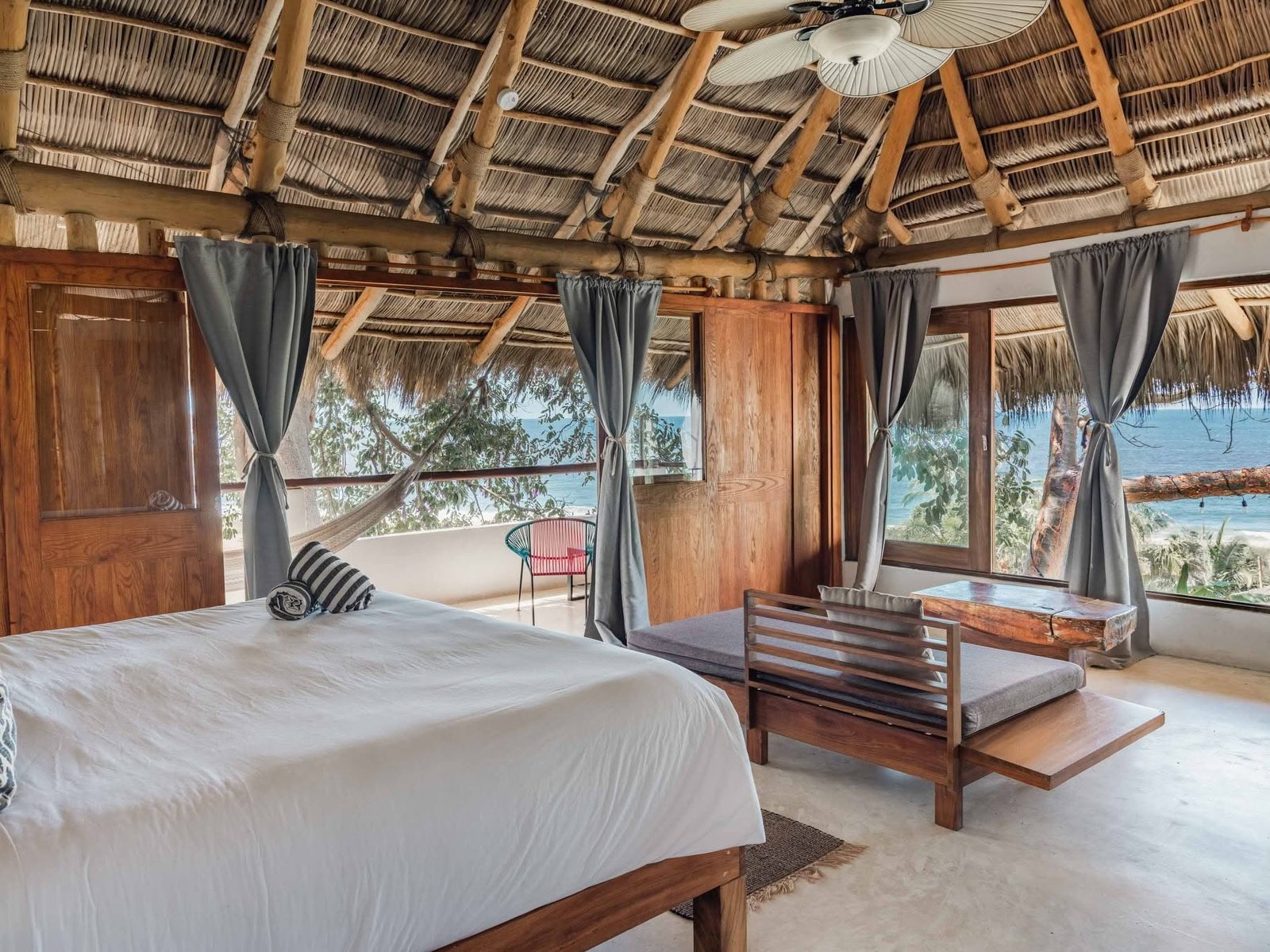 Suite con cama king y ventanal abierto a la bahía bajo techo de palapa
